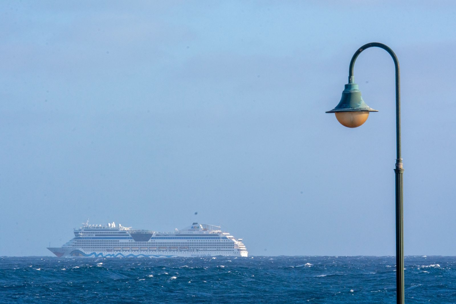 Cruise ship on the open Pacific horizon seen past a seaside street lamp, the one-way voyages behind repositioning cruise deals